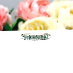 A sterling silver 5-stone band set with 3mm round sparkling aquamarine blue moissanites. Displayed on a white surface with a floral background.