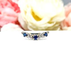 A sterling silver curved 5-stone band featuring alternating round lab blue sapphires and moissanites. The band has a classic design with the gemstones set in a row with three sapphires and two moissanites. Displayed on a white surface with a pastel floral background.