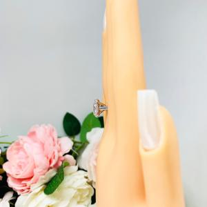 Mannequin hand with long white shimmery nails, displayed against a soft white-gray background with pink and white floral accents. The hand is wearing a sterling silver solitaire engagement ring set with a 12x8mm elongated oval cubic zirconia. The ring features split prongs and a cathedral shank.