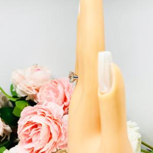 Mannequin hand with long white shimmery nails, displayed against a soft white-gray background with pink and white floral accents. The hand is wearing a near colorless (G/H/I color) round brilliant cut cubic zirconia solitaire ring in a split prong cathedral setting. The ring is made of sterling silver and features a sleek, minimalist design. The setting is simple yet elegant, allowing the center stone to take center stage.
