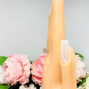 Mannequin hand with long white shimmery nails, displayed against a soft white-gray background with pink and white floral accents. The hand is wearing a near colorless (G/H/I color) round brilliant cut cubic zirconia solitaire ring in a split prong cathedral setting. The ring is made of sterling silver and features a sleek, minimalist design. The setting is simple yet elegant, allowing the center stone to take center stage.