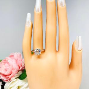 Mannequin hand with long white shimmery nails, displayed against a soft white-gray background with pink and white floral accents. The hand is wearing a near colorless (G/H/I color) round brilliant cut cubic zirconia solitaire ring in a split prong cathedral setting. The ring is made of sterling silver and features a sleek, minimalist design. The setting is simple yet elegant, allowing the center stone to take center stage.