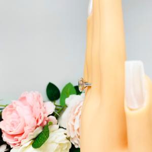 Mannequin hand with long white shimmery nails, displayed against a soft white-gray background with pink and white floral accents. The hand is wearing a sterling silver solitaire engagement ring set with a 12x8mm elongated oval near colorless cubic zirconia. The ring features split prongs and a cathedral shank.