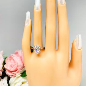 Mannequin hand with long white shimmery nails, displayed against a soft white-gray background with pink and white floral accents. The hand is wearing a sterling silver solitaire engagement ring set with a 12x8mm elongated oval near colorless cubic zirconia. The ring features split prongs and a cathedral shank.
