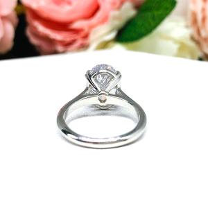 Sterling silver solitaire engagement ring set with a 12x8mm elongated oval cubic zirconia. The ring features split prongs and a cathedral shank. It is resting on a white surface with soft-focus pink and white flowers in the background.