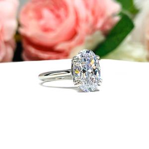 Sterling silver solitaire engagement ring set with a 12x8mm elongated oval cubic zirconia. The ring features split prongs and a cathedral shank. It is resting on a white surface with soft-focus pink and white flowers in the background.