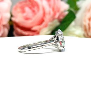 Sterling silver solitaire engagement ring set with a 12x8mm elongated oval cubic zirconia. The ring features split prongs and a cathedral shank. It is resting on a white surface with soft-focus pink and white flowers in the background.