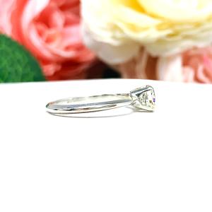 Sterling silver engagement ring set with an 6mm brilliant cushion near colorless cubic zirconia. The ring is resting on a white surface with a background of soft-focus pink and white flowers.