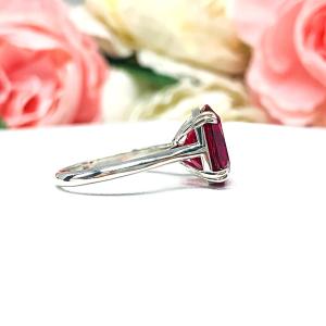 Sterling silver ring featuring a vivid elongated cushion-cut lab created ruby held in a split-prong setting, with soft-focus pink and white flowers in the background and “SJ” initials subtly visible in the corner.