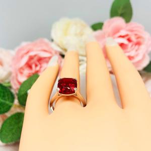 Mannequin hand with long white shimmery nails, displayed against a soft white-gray background with pink and white floral accents. The hand is wearing a sterling silver ring featuring a vivid elongated cushion-cut lab created ruby held in a split-prong setting.