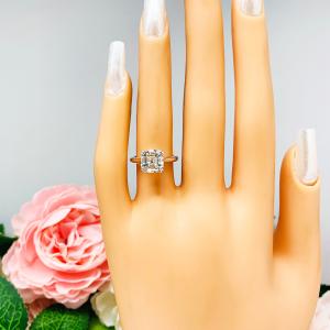 Mannequin hand with long white shimmery nails, displayed against a soft white-gray background with pink and white floral accents. The hand is wearing a sterling silver 8mm near colorless (G/H color) asscher cut cubic zirconia solitaire engagement ring. The ring is resting on a white surface with a background of pink and white flowers.
