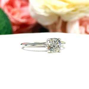 Sterling silver engagement ring set with an 6mm brilliant cushion near colorless cubic zirconia. The ring is resting on a white surface with a background of soft-focus pink and white flowers.
