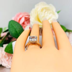 Mannequin hand with long white shimmery nails, displayed against a soft white-gray background with pink and white floral accents. The hand is wearing a sterling silver 9mm near colorless (G/H color) asscher cut cubic zirconia solitaire engagement ring.