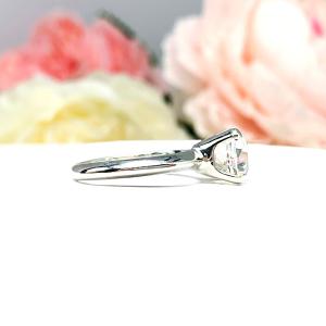 Sterling silver engagement ring set with an 8mm old mine cut near colorless cubic zirconia. The ring is resting on a white surface with a background of soft-focus pink and white flowers.