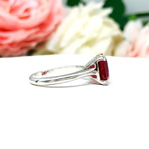 Sterling silver ring with a cushion cut lab created deep red ruby set in a split-prong setting set against a white surface with soft-focus pink and white flowers in the background.