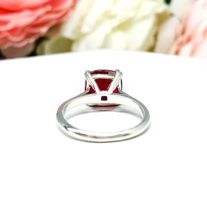 Sterling silver ring with a cushion cut lab created deep red ruby set in a split-prong setting set against a white surface with soft-focus pink and white flowers in the background.
