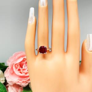 Mannequin hand with long white shimmery nails, displayed against a soft white-gray background with pink and white floral accents. The hand is wearing a sterling silver ring with a cushion cut lab created deep red ruby set in a split-prong setting.