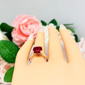 Mannequin hand with long white shimmery nails, displayed against a soft white-gray background with pink and white floral accents. The hand is wearing a sterling silver ring with a cushion cut lab created deep red ruby set in a split-prong setting.