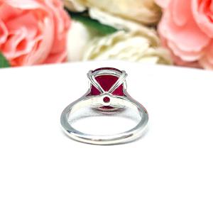 Sterling silver ring featuring a vivid elongated cushion-cut lab created ruby held in a split-prong setting, with soft-focus pink and white flowers in the background and “SJ” initials subtly visible in the corner.