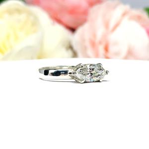 A 6-prong sterling silver, horizontal east west set engagement ring featuring a precision-faceted near colorless (G/H color) marquise cubic zirconia.  Displayed on a white surface with soft pastel floral background.