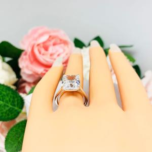 Mannequin hand with long white shimmery nails, displayed against a soft white-gray background with pink and white floral accents. The hand is wearing an elegant sterling silver cathedral engagement ring set with a large radiant cut cubic zirconia.