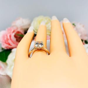 A display hand with long white fingernails standing up and with the top of the hand facing forward, displayed against a soft-focus background of pink and white flowers. The display hand is wearing sterling silver cathedral-style engagement ring featuring an 8mm near colorless cushion cut cubic zirconia center stone.