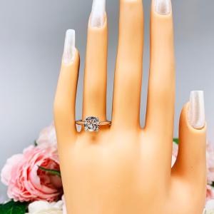 A display hand with long white fingernails standing up and with the top of the hand facing forward, displayed against a soft-focus background of pink and white flowers. The display hand is wearing sterling silver cathedral-style engagement ring featuring an 8mm near colorless cushion cut cubic zirconia center stone. The ring is shown facing forward, showcasing the brilliant faceting of the center stone and split prongs.