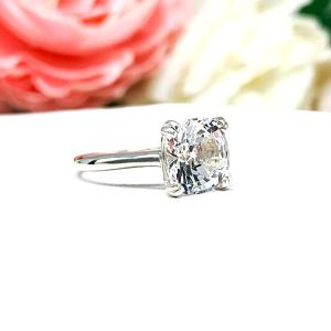 Sterling silver engagement ring with a sparkling cushion cut lab created white sapphire set in a split-prong setting set against a white surface with soft-focus pink and white flowers in the background.