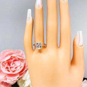 Mannequin hand with long white shimmery nails, displayed against a soft white-gray background with pink and white floral accents. The hand is wearing a sterling silver ring with a sparkling cushion cut lab created white sapphire set in a split-prong setting.