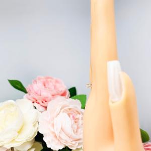 Mannequin hand with long white shimmery nails, displayed against a soft white-gray background with pink and white floral accents. The hand is wearing a dainty 4-prong sterling silver, horizontal east west set engagement ring featuring a precision-faceted marquise cubic zirconia.