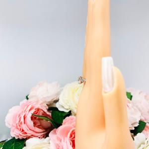 A display hand with long white fingernails standing up and with the top of the hand facing forward, displayed against a soft-focus background of pink and white flowers. The display hand is wearing sterling silver cathedral-style engagement ring featuring an 8mm near colorless cushion cut cubic zirconia center stone.