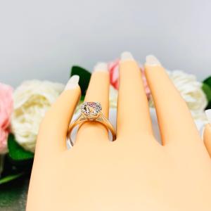A display hand with long white fingernails standing up and with the top of the hand facing forward, displayed against a soft-focus background of pink and white flowers. The display hand is wearing a 9mm (3 ct) old European cut near-colorless CZ solitaire engagement ring. Set in a split prong cathedral-style mount, the ring gleams with vintage-inspired elegance.