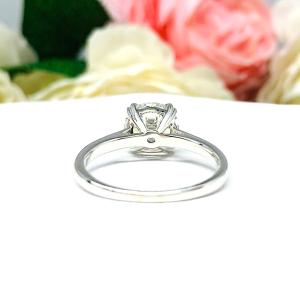 A romantic close-up of a 9mm (3 ct) old European cut near-colorless CZ solitaire engagement ring. Set in a sterling silver split prong cathedral-style mount, the ring gleams with vintage-inspired elegance. Soft pink and white flowers blur in the background, adding a dreamy, bridal ambiance.