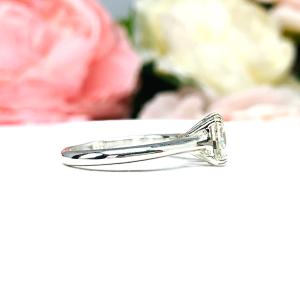 Sterling silver split prong cathedral engagement ring set with an 7.5mm near colorless old mine cut cubic zirconia. The ring is resting on a white surface with a background of pink and white flowers.