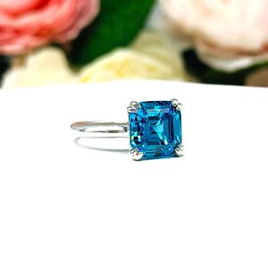 Sterling silver 8mm teal blue asscher cut cubic zirconia solitaire ring. The ring is resting on a white surface with a background of pink and white flowers.