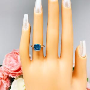 Mannequin hand with long white shimmery nails, displayed against a soft white-gray background with pink and white floral accents. The hand is wearing a sterling silver 8mm teal blue asscher cut cubic zirconia solitaire ring.
