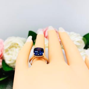 Mannequin hand with long white shimmery nails, displayed against a soft white-gray background with pink and white floral accents. The hand is wearing a sterling silver ring featuring a vivid elongated cushion-cut lab created blue sapphire held in a split-prong setting.