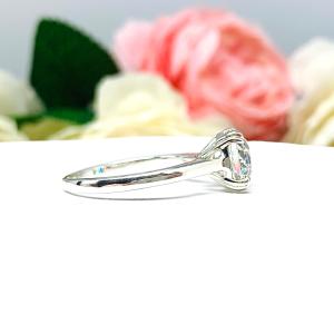 A romantic close-up of a 9mm (3 ct) old European cut near-colorless CZ solitaire engagement ring. Set in a sterling silver split prong cathedral-style mount, the ring gleams with vintage-inspired elegance. Soft pink and white flowers blur in the background, adding a dreamy, bridal ambiance.