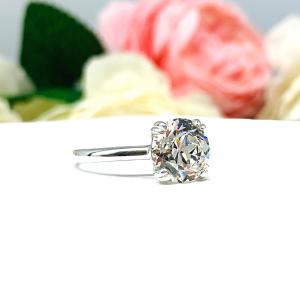 A romantic close-up of a 9mm (3 ct) old European cut near-colorless CZ solitaire engagement ring. Set in a sterling silver split prong cathedral-style mount, the ring gleams with vintage-inspired elegance. Soft pink and white flowers blur in the background, adding a dreamy, bridal ambiance.