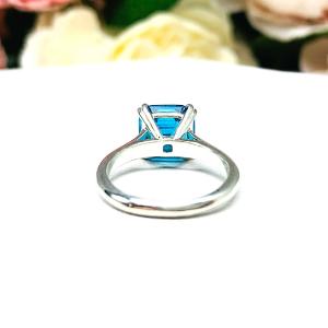 Sterling silver 8mm teal blue asscher cut cubic zirconia solitaire ring. The ring is resting on a white surface with a background of pink and white flowers.