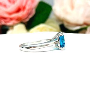 Sterling silver 8mm teal blue asscher cut cubic zirconia solitaire ring. The ring is resting on a white surface with a background of pink and white flowers.