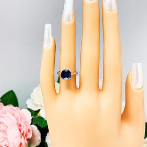 A display hand with long white fingernails standing up and with the top of the hand facing forward, displayed against a soft-focus background of pink and white flowers. The display hand is wearing a sterling silver cathedral-style ring featuring an 8mm cushion cut lab created blue sapphire center stone.