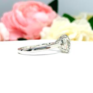 A romantic close-up of a 10mm (4 ct) old European cut near-colorless CZ solitaire engagement ring. Set in a sterling silver split prong cathedral-style mount, the ring gleams with vintage-inspired elegance. Soft pink and white flowers blur in the background, adding a dreamy, bridal ambiance.