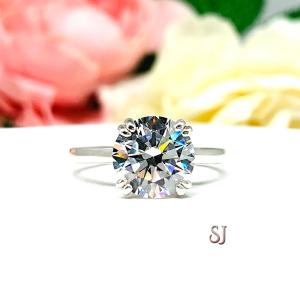 Close-up view of a colorless round brilliant cut cubic zirconia solitaire ring in a split prong cathedral setting. The ring is made of sterling silver and features a sleek, minimalist design. The setting is simple yet elegant, allowing the center stone to take center stage. The ring is displayed against a soft, blurred background of pastel-colored flowers, creating a romantic and delicate ambiance.