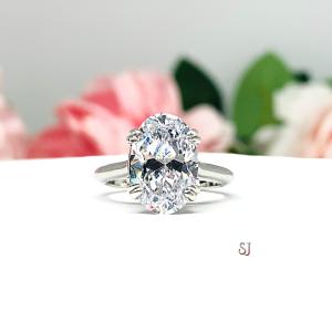 Sterling silver solitaire engagement ring set with a 12x8mm elongated oval cubic zirconia. The ring features split prongs and a cathedral shank. It is resting on a white surface with soft-focus pink and white flowers in the background.