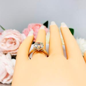 A display hand with long white fingernails laying down and with the top of the hand facing up, displayed against a soft-focus background of pink and white flowers.  The display hand is wearing a 10mm (4 ct) old European cut near-colorless CZ solitaire engagement ring. Set in a split prong cathedral-style mount, the ring gleams with vintage-inspired elegance.