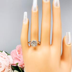 A display hand with long white fingernails standing up and with the top of the hand facing forward, displayed against a soft-focus background of pink and white flowers. The display hand is wearing a 10mm (4 ct) old European cut near-colorless CZ solitaire engagement ring. Set in a split prong cathedral-style mount, the ring gleams with vintage-inspired elegance.