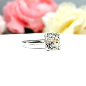 A sterling silver split-prong cathedral engagement ring set with a near colorless old mine cut cubic zirconia. The ring is set against a white background with a blurred image of pink and white flowers in the background.