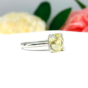 Sterling silver split prong cathedral engagement ring set with an 8mm light yellow old mine cut cubic zirconia. The ring is resting on a white surface with a background of pink and white flowers.