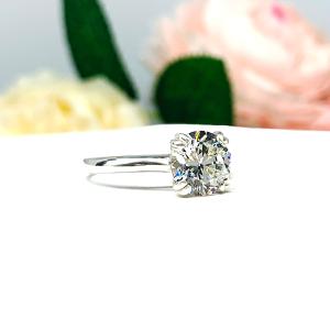 Close-up view of a near colorless (G/H color) round brilliant cut cubic zirconia solitaire ring in a split prong cathedral setting. The ring is made of sterling silver and features a sleek, minimalist design. The setting is simple yet elegant, allowing the center stone to take center stage. The ring is displayed against a soft, blurred background of pastel-colored flowers, creating a romantic and delicate ambiance.